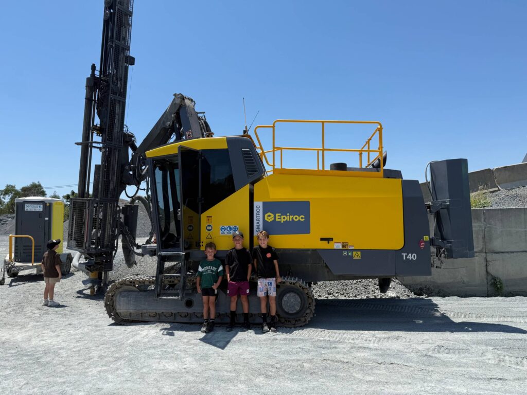 Three young boys standing proudly in front of a massive yellow and grey Epiroc Smartroc T40 surface drill rig on a gravel worksite.