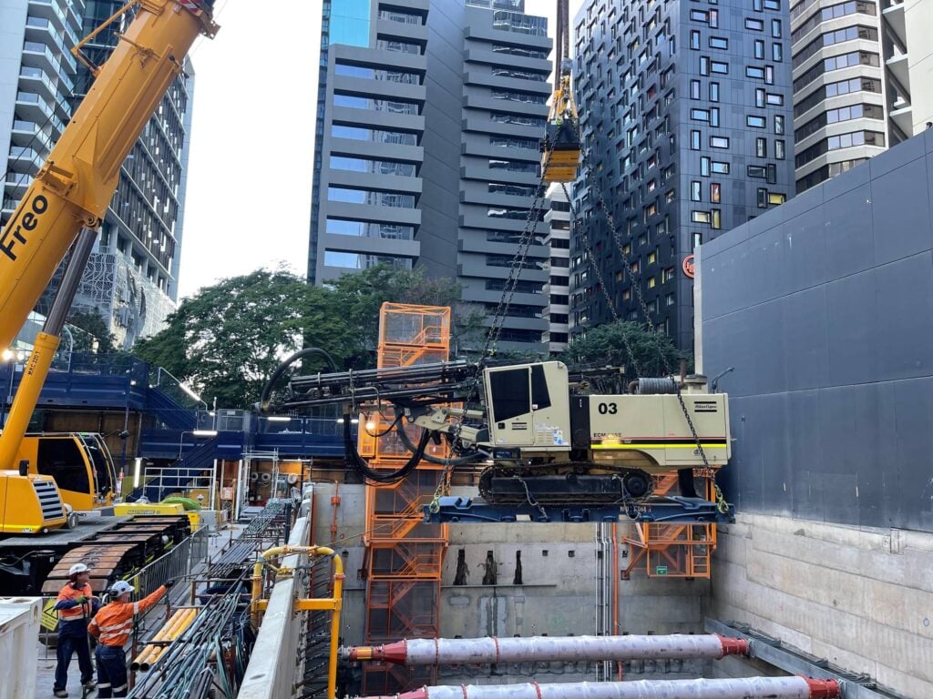 A large yellow crane lowers a tan Atlas Copco drill rig into a deep construction pit in a dense urban area