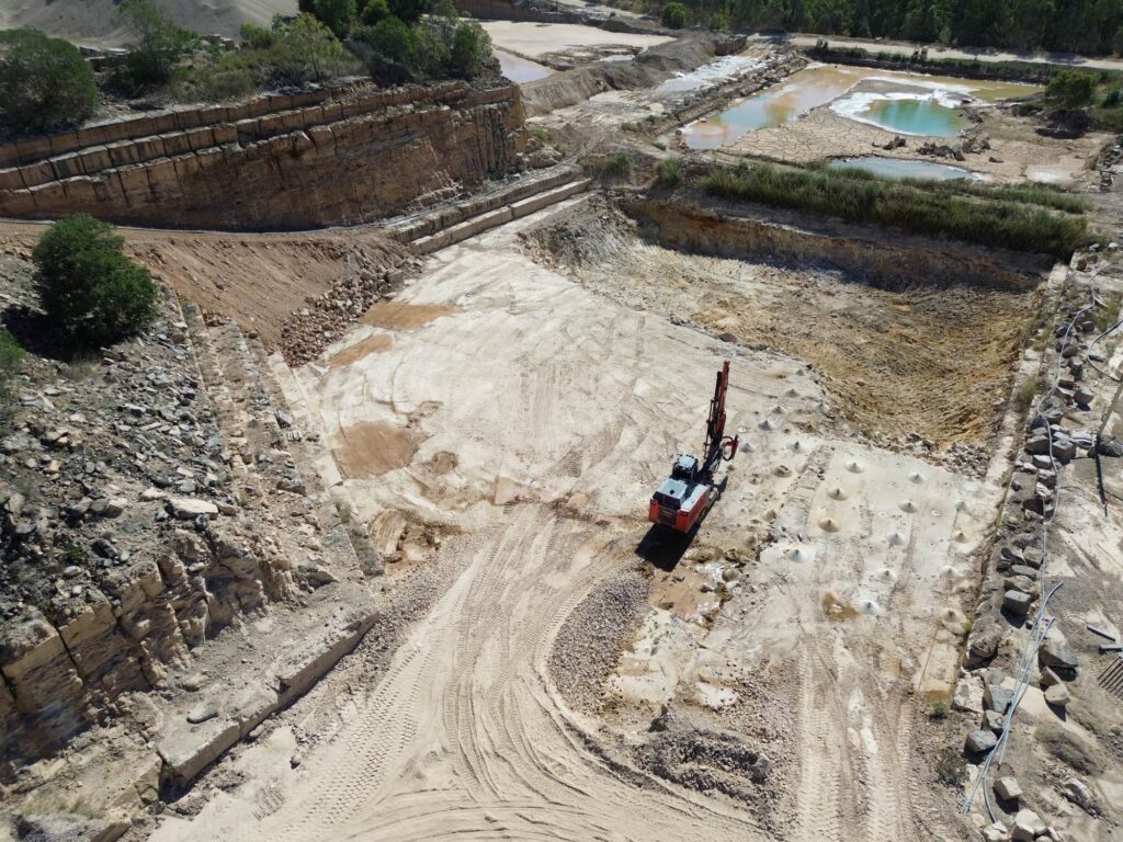 A wide, terraced quarry with a red drill rig working on a flat bench