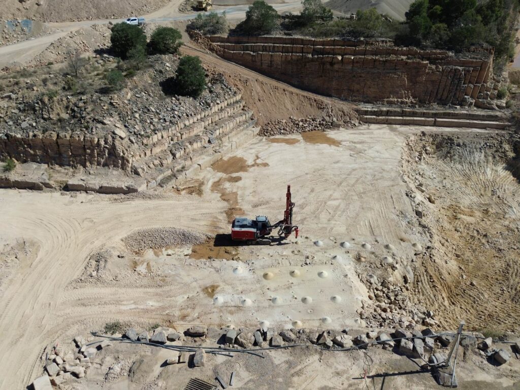 A red and white surface drill rig working on a light-coloured bench in a terraced stone quarry